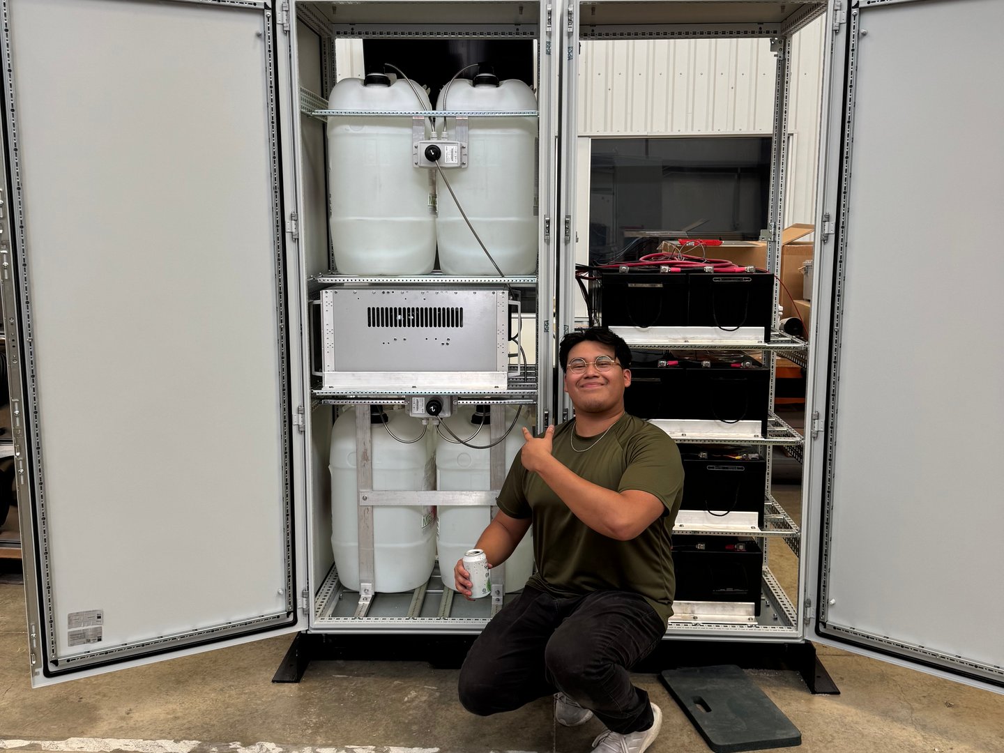 A team member crouching with a thumbs up in front of an open power enclosure full of fuel tanks and batteries